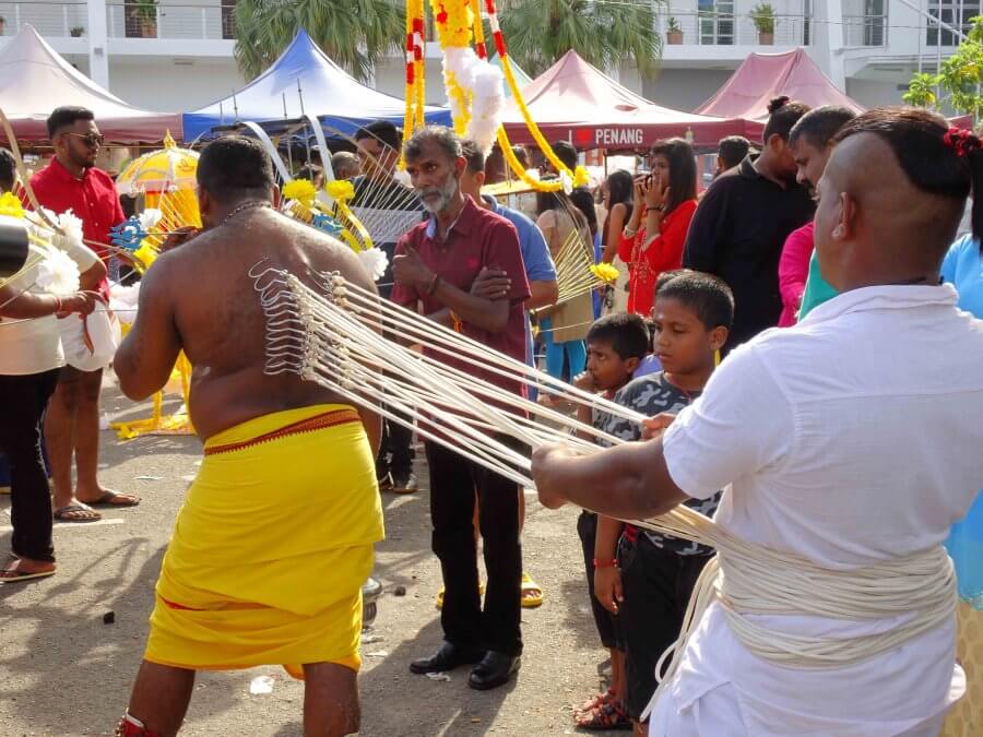 A man looking on at one of the devotees who has a spear through his face and ropes being pulled from his back. Thaipusam devotees