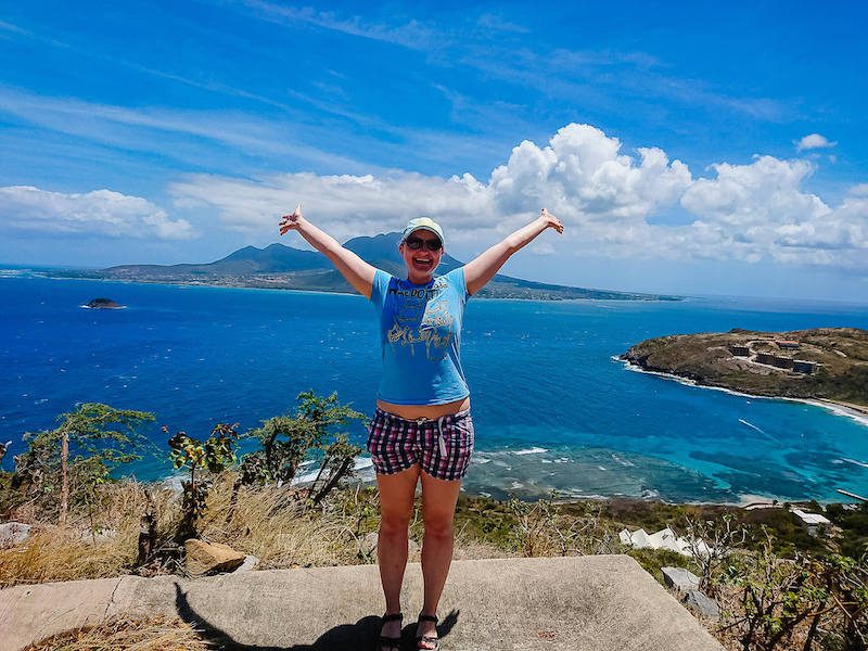 Stephanie with arms outstretched in front of blue ocean and mountains of St Kitts. Reasons to move to another country.