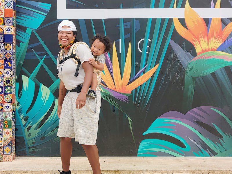 happy momma carrying her son on her back in front of a mural of tropical flowers