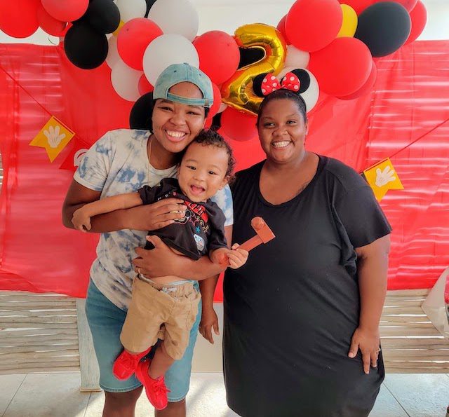 two women with a baby in front of balloons and a red background