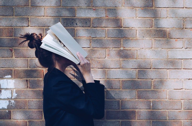 girl with book on face in front of brink wall