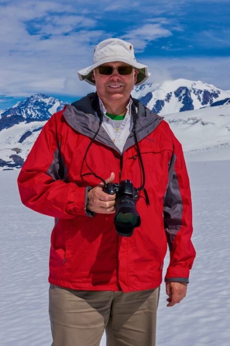 man with sunglasses, hat and red jacket standing with camera in snowy mountains