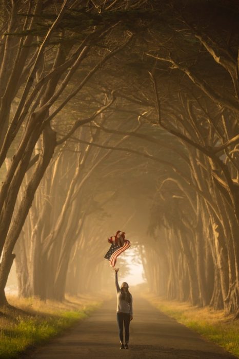 girl standing on a tree lined street on a foggy day holding the American flag